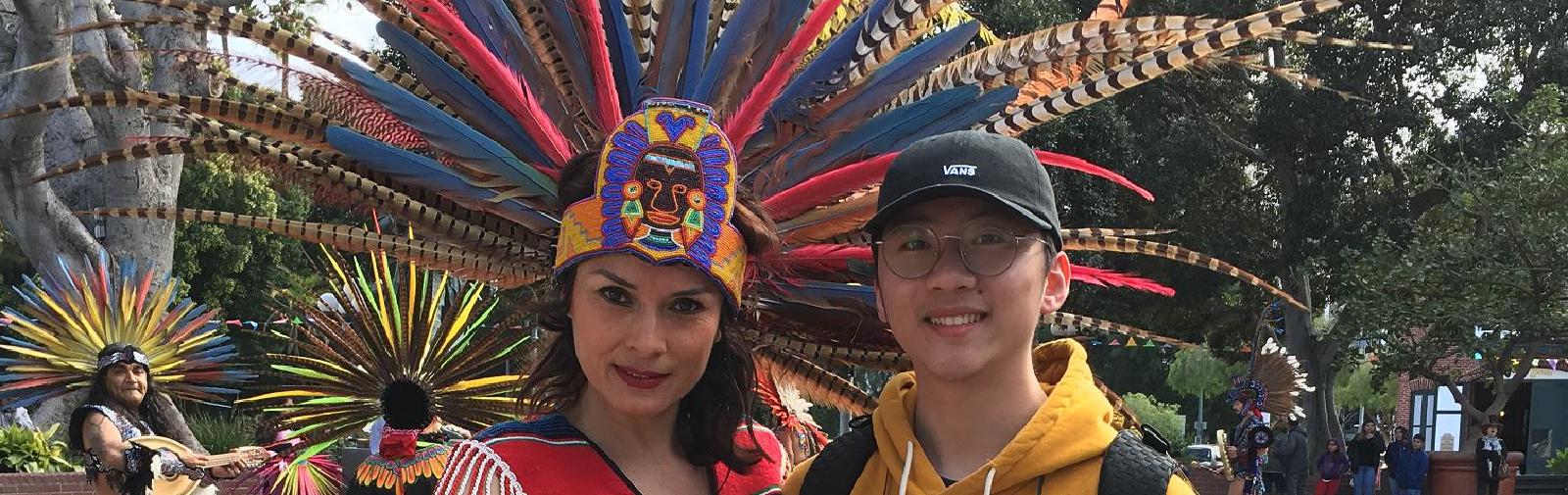 A woman with an elaborate costume with colorful feathers posing with a tour goer.